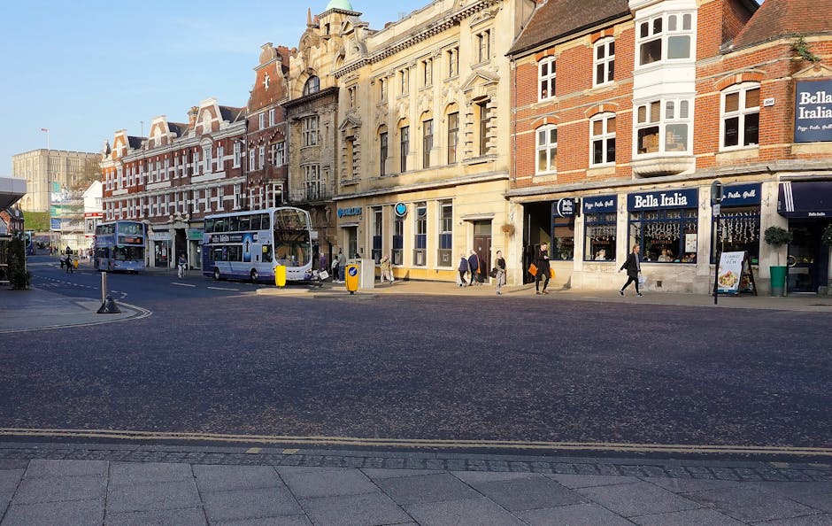 A busy street scene featuring a row of multi-story buildings with traditional brick and stone facades, large windows, and shopfronts including a restaurant named Bella Italia. There are several pedest